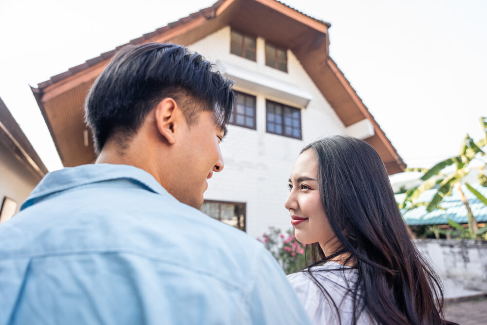 Asian young happy new marriage couple moving to new house together. Attractive romantic man and woman feeling happy and excited, standing in front of new home after buying together with estate agent.
