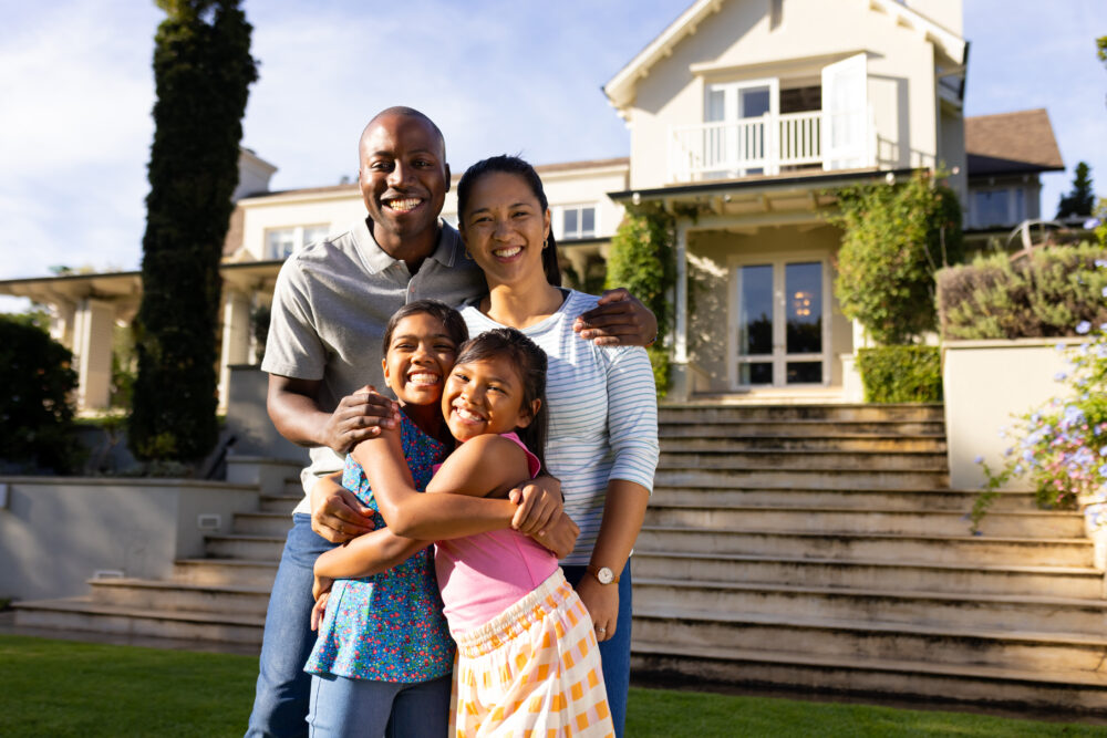 Outdoors, diverse family of four smiling and embracing in front of their home. The background shows a beautiful, well-maintained house with a lush garden and stone steps, unaltered